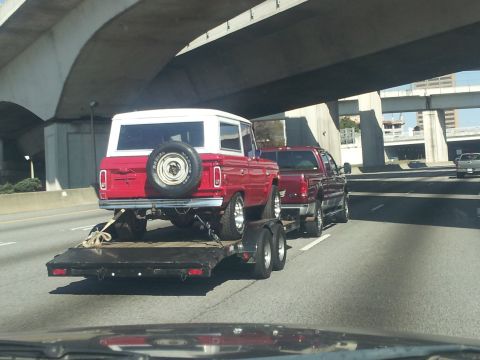 1986 Ford Bronco - A red and white SUV, likely a 1978 Ford Bronco, is transported on a trailer behind a pickup truck on the highway.