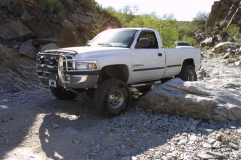 1994 Ford Bronco - A white 1994 Dodge Ram pickup truck, modified for off-roading, is shown traversing a rocky, uneven path with another vehicle visible in the…