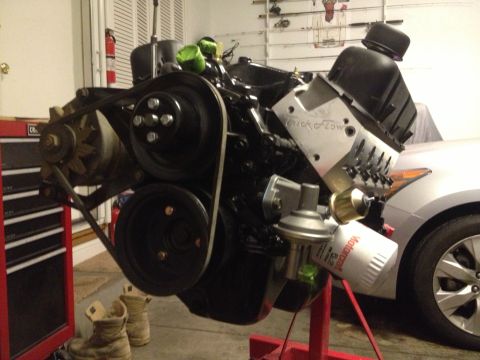 1965 Ford Mustang - A V8 engine is mounted on a stand, with a fuel pump and oil filter attached. A silver car is partially visible in the background.