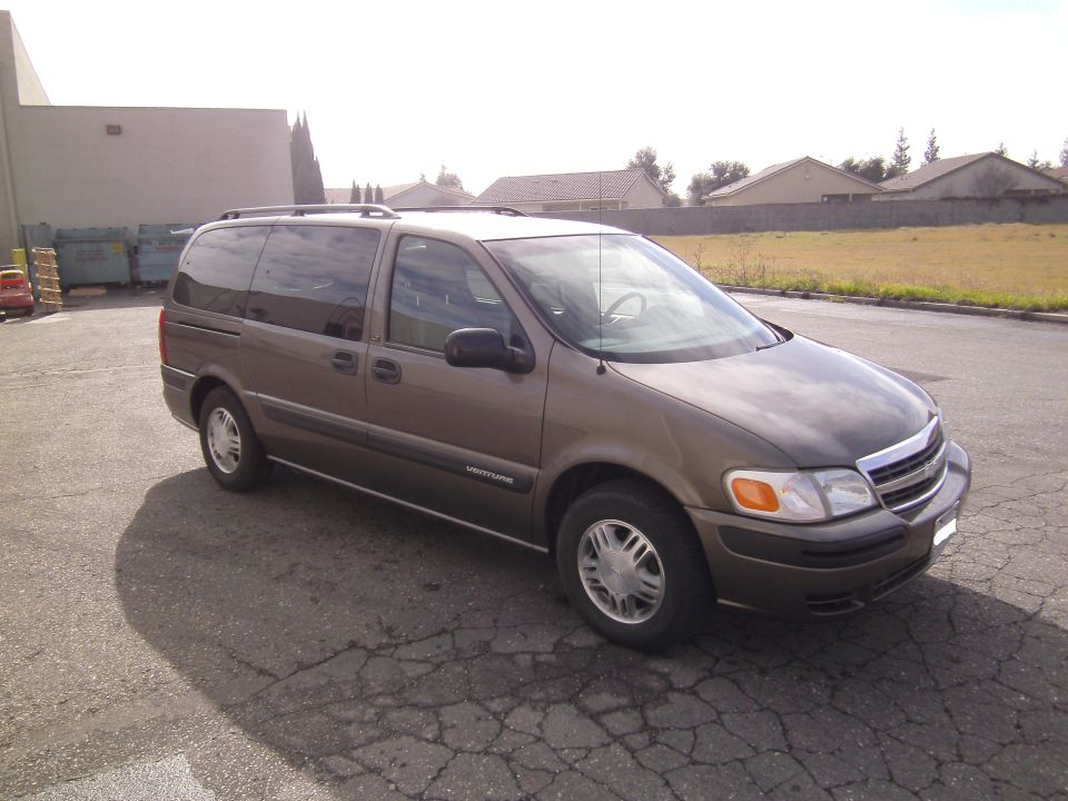 Brown minivan parked on cracked asphalt, with a building and some houses in the background