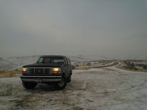 1992 Ford Bronco - A black Ford Bronco is parked on a snowy dirt road, with a winding road disappearing into the hazy hills.