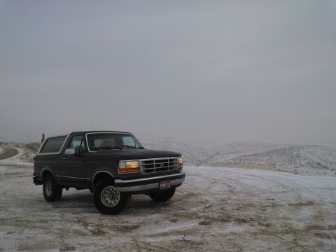 1992 Ford Bronco - A dark 1992 Ford Bronco with a white top is parked on a snowy road with hills in the background.