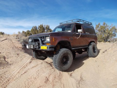 1986 Ford Bronco II - Modified brown Ford Bronco II navigates a dirt track under a clear blue sky.