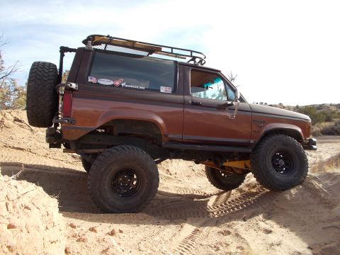 1986 Ford Bronco II - This lifted 1986 Ford Bronco II navigates a sandy incline, equipped with large off-road tires and a roof rack.
