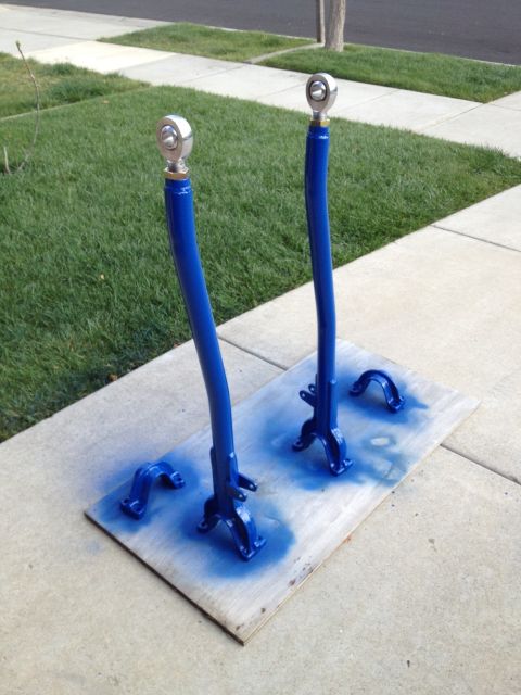 1971 Ford Bronco - Two freshly painted blue custom radius arms with silver ball joints are laid out on a wooden board on a sidewalk.