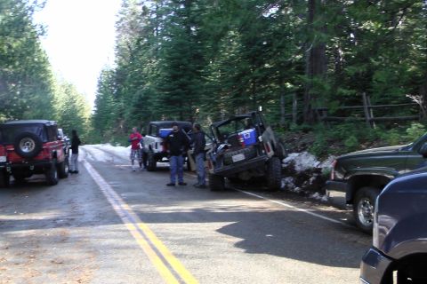 1971 Ford Bronco - A group of off-road vehicles are stopped on a road with snow on the shoulder, with several people gathered nearby.