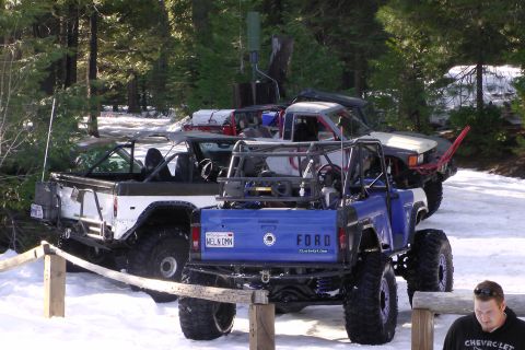 1971 Ford Bronco - Two modified Ford Broncos are parked in a snowy wooded area, with the blue Bronco in the foreground showing its "Ford" tailgate.