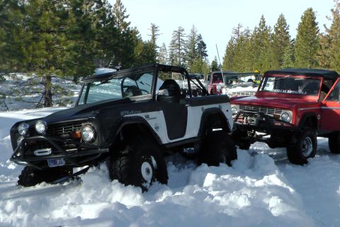 1971 Ford Bronco - Multiple Ford Broncos navigate a snowy terrain, with two prominent vehicles partially buried in snow.