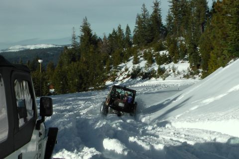 1971 Ford Bronco - Two Jeep Wranglers navigate a snow-covered road with pine trees and mountains in the background.