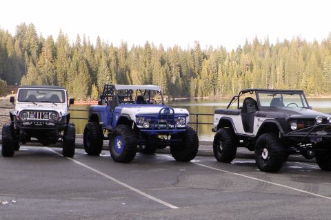 1971 Ford Bronco - Three lifted off-road vehicles are parked side-by-side in a paved lot next to a body of water.