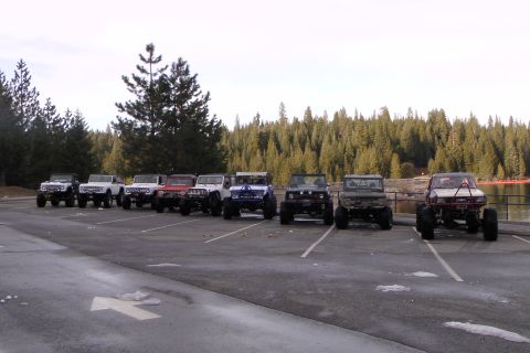 1971 Ford Bronco - A row of classic Ford Broncos are parked in a lot, with a dense forest and calm lake visible in the background.