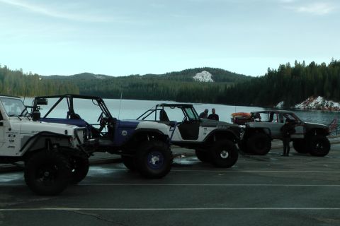 1971 Ford Bronco - Modified off-road vehicles are lined up next to a body of water with a forested hillside behind them.