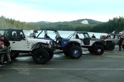 1971 Ford Bronco - Modified off-road vehicles are lined up next to a body of water on a cloudy day.