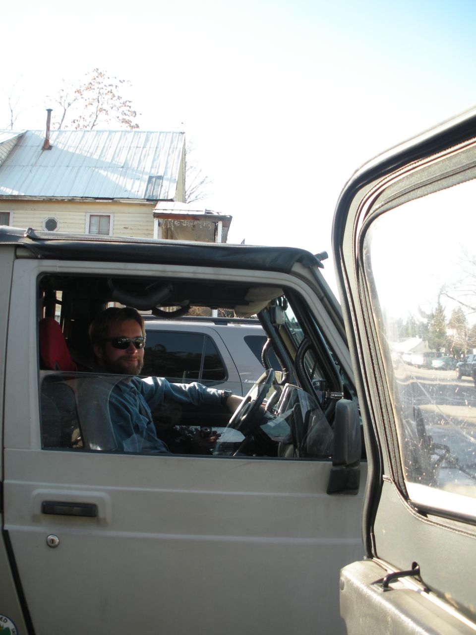 Man wearing sunglasses and a blue shirt sits in the driver's seat of a silver SUV. Another SUV is visible behind it