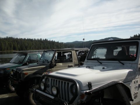 1971 Ford Bronco - Several off-road vehicles are parked in a line near a body of water with trees and mountains in the background.