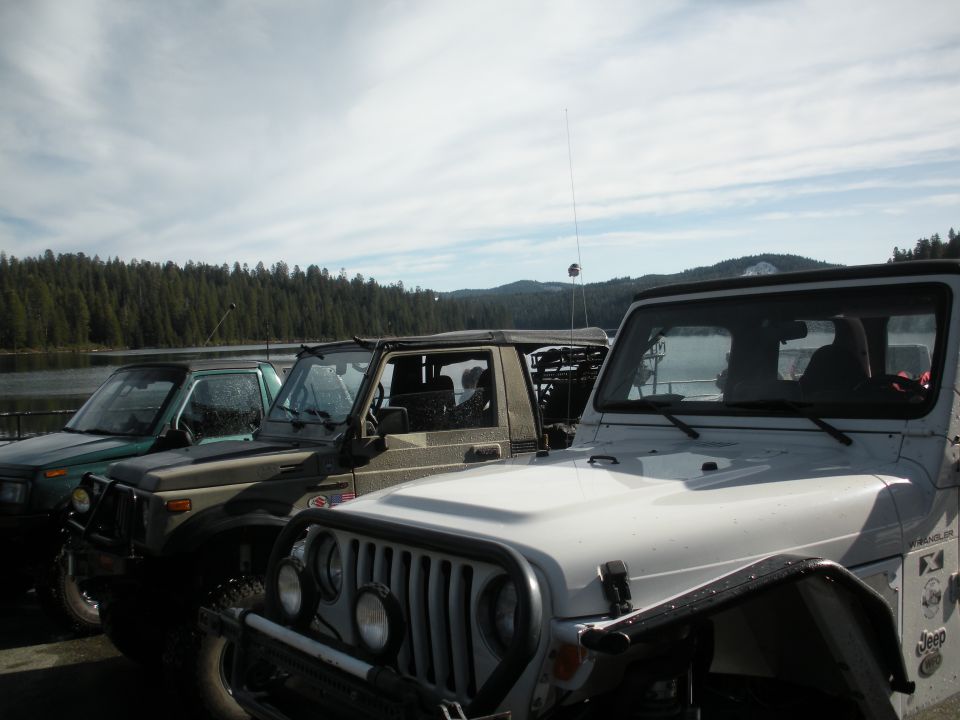 White Jeep Wrangler in foreground, with other SUVs parked in a line next to a lake and forest