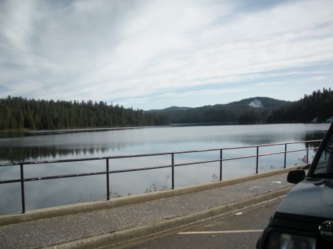 1971 Ford Bronco - A dark SUV is parked next to a calm lake surrounded by a dense forest and distant hills.