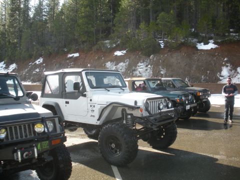 1971 Ford Bronco - Several off-road vehicles, including a white Jeep Wrangler, are parked in a lot with snow on the surrounding hills.