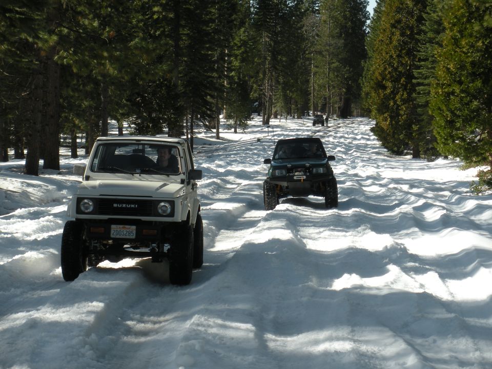 Two Suzuki Samurai SUVs driving on a snow-covered road through a pine forest. The lead vehicle is white, the second is dark green