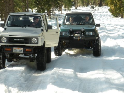 1971 Ford Bronco - Two off-road vehicles navigate a snow-covered trail surrounded by pine trees.