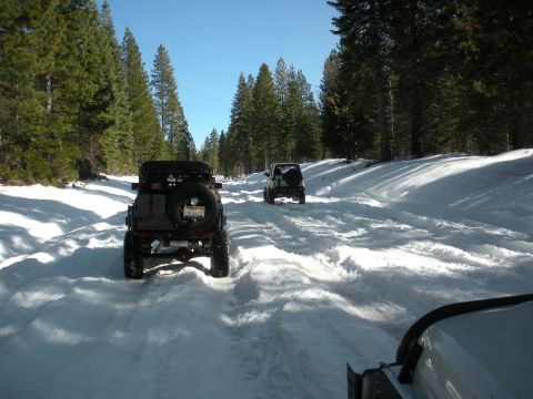 1971 Ford Bronco - Two SUVs navigate a snow-covered forest road under a clear blue sky.