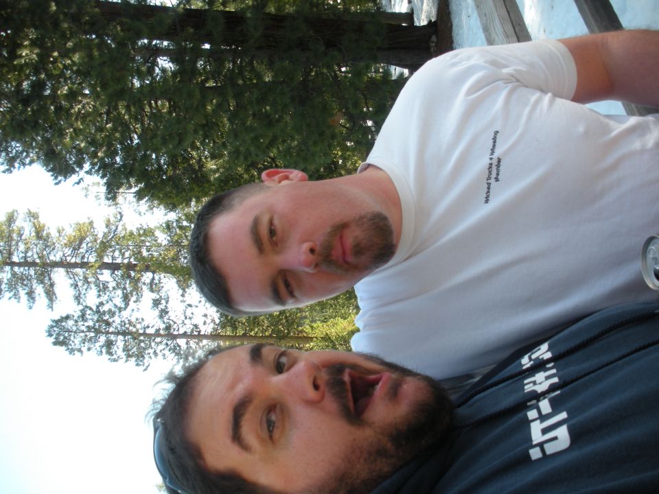 Two men pose for a selfie outdoors with trees in the background. One man wears a white t-shirt with text