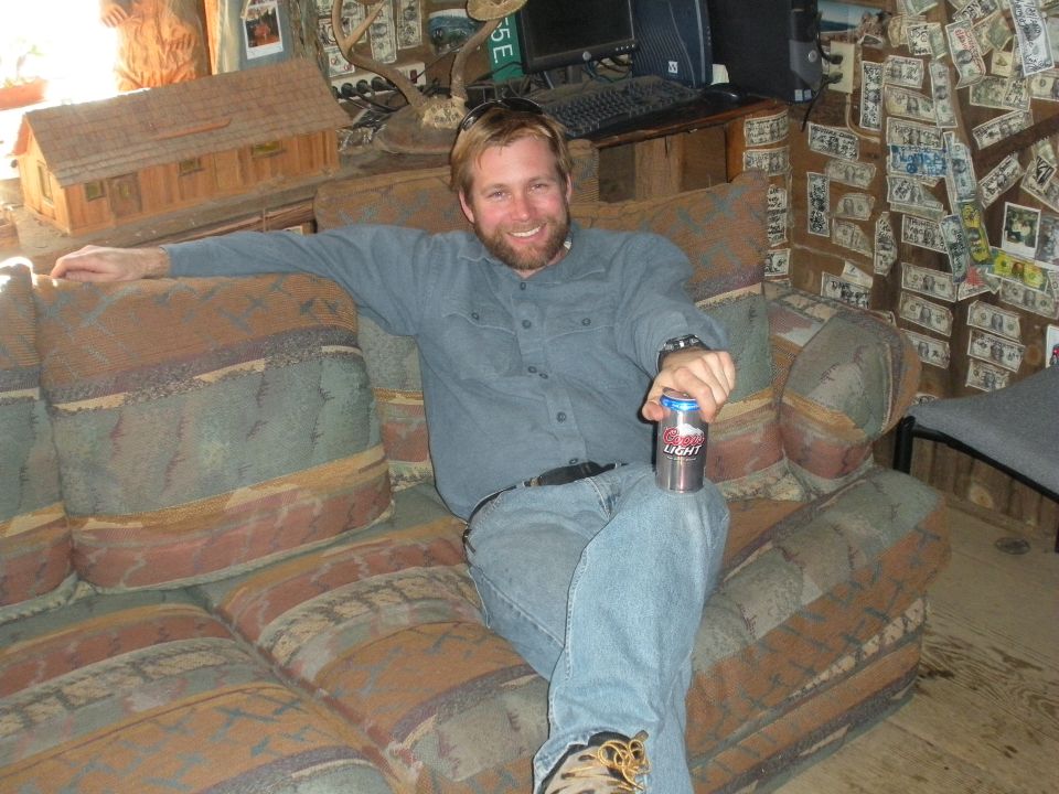 Guy sitting on a couch holding a beer, room decorated with money and wooden models