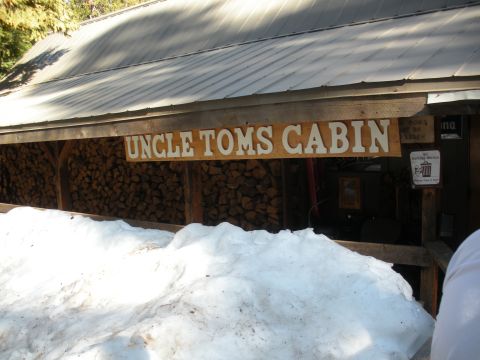 1971 Ford Bronco - A wooden sign reads "Uncle Toms Cabin" above a pile of firewood, with snow in the foreground.