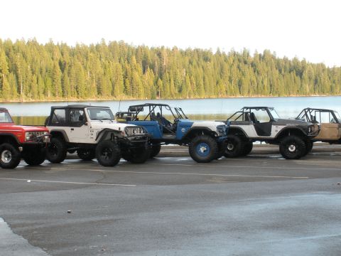1971 Ford Bronco - A row of customized off-road vehicles are parked by a lake, with a dense forest visible behind them.