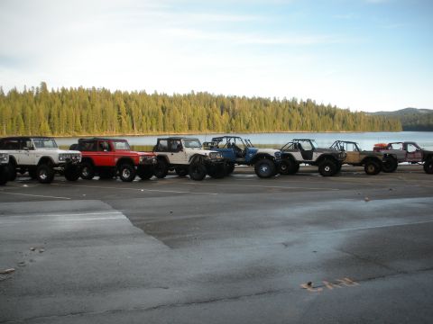 1971 Ford Bronco - A fleet of modified off-road vehicles, including a red Ford Bronco, are lined up near a body of water with a forest in the background.