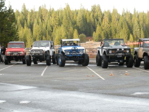 1971 Ford Bronco - A line of modified off-road vehicles, including a red 1971 Ford Bronco, are parked in front of a forest.