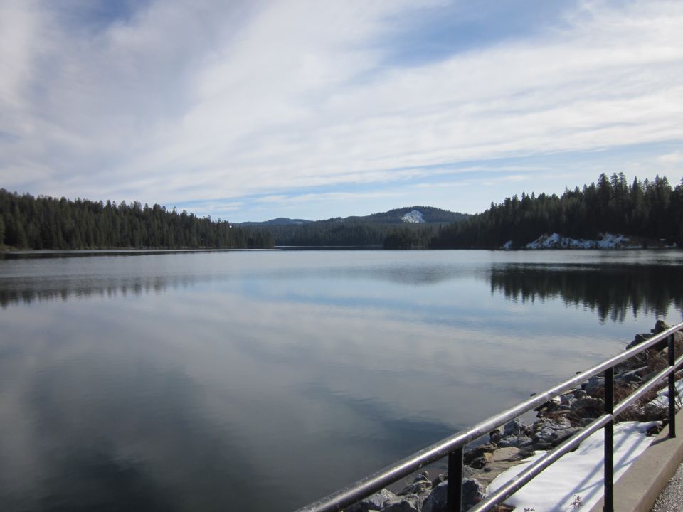 Calm lake reflects cloudy sky, with dense pine forest on the far shore and a snowy patch on a distant hill. A metal railing is in the foreground
