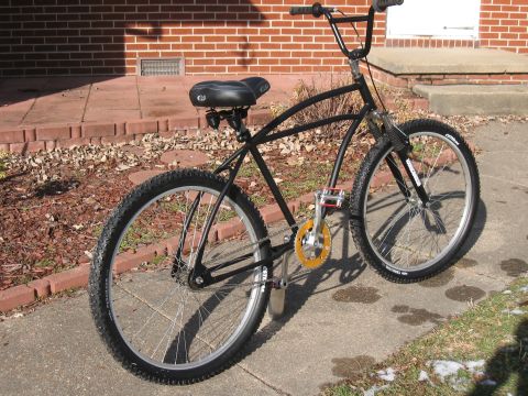1994 Ford Bronco - A black cruiser bicycle with wide tires and yellow pedals is parked on a concrete sidewalk.
