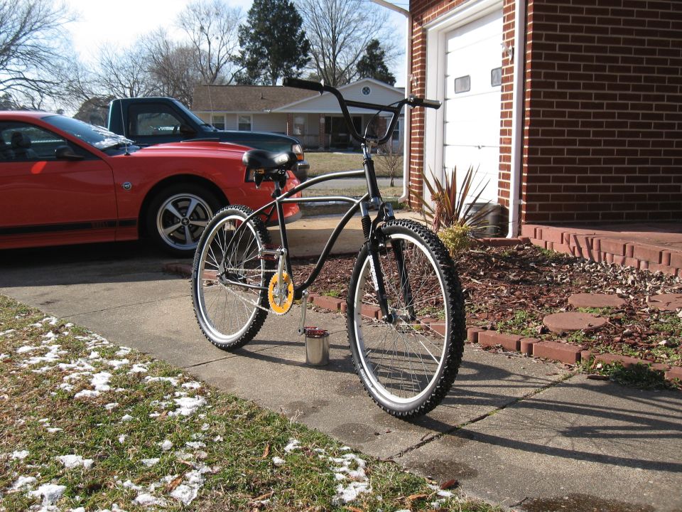Black cruiser bicycle parked on sidewalk in front of a red car and a brick house with a garage