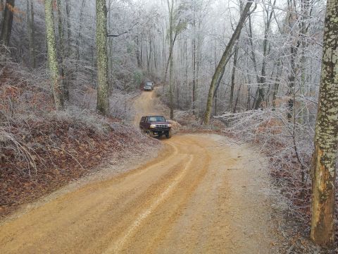1986 Ford Bronco - Two SUVs navigate a winding dirt road through a forest with icy branches and trees.