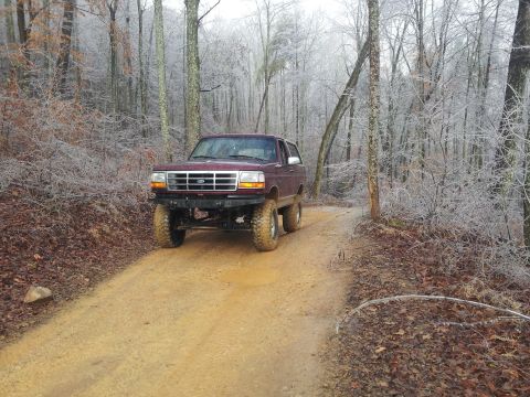 1986 Ford Bronco - Maroon Ford Bronco driving on a dirt road through a forest with icy trees.