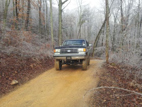 1986 Ford Bronco - A lifted Ford truck drives down a muddy, wooded trail on a cold, icy day.