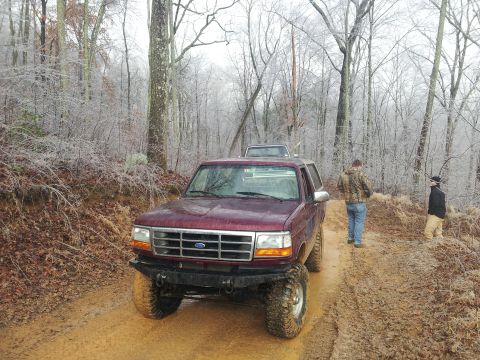 1986 Ford Bronco - A maroon SUV navigates a muddy, wooded trail with another vehicle following behind.