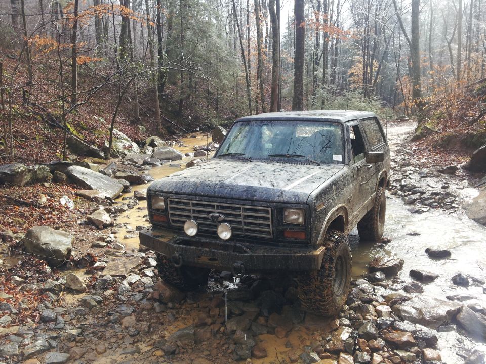 Dark blue Ford Bronco driving through a rocky stream bed in a forest