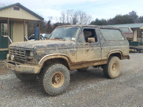 1986 Ford Bronco - A heavily muddied 1980s Ford Bronco sits parked on a gravel lot near a small building.