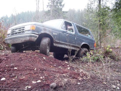 1987 Ford Bronco - A dark blue 1987 Ford Bronco navigates a challenging off-road terrain with muddy tires and a tilted body.