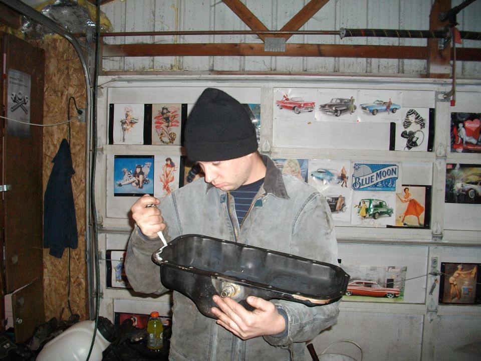 Man in garage applying sealant to a black oil pan. Garage door has vintage car pictures and a Blue Moon sign