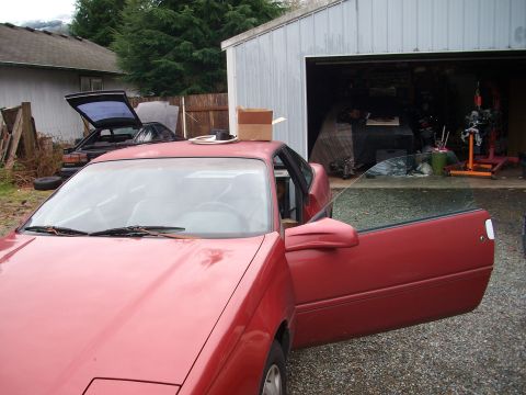 1989 Ford Probe - A red coupe sits in a driveway with its door ajar, parked near a garage and another vehicle.