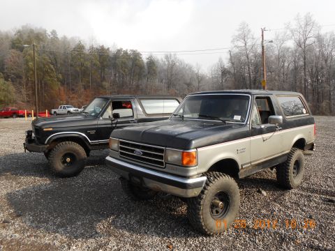 1986 Ford Bronco - Two Ford Broncos are parked on a gravel lot with trees in the background. The front Bronco is a two-tone model.