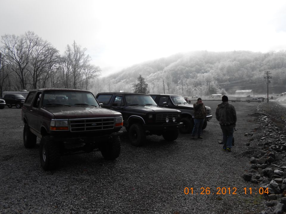 Three SUVs parked in a gravel lot with trees and a misty mountain in the background. The first SUV is a maroon Ford Bronco. Two men stand near the other SUVs