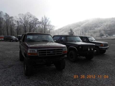 1986 Ford Bronco - Three Ford Broncos are parked in a gravel lot with trees and a misty hillside in the background.