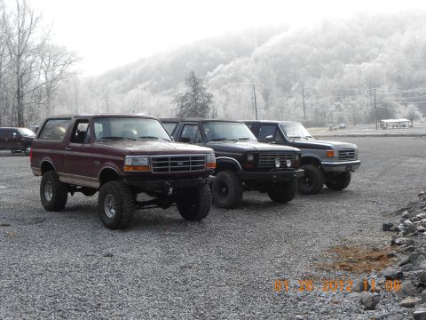 1986 Ford Bronco - Three lifted trucks are parked in a gravel lot on a foggy day, with a line of trees and hills in the distance.