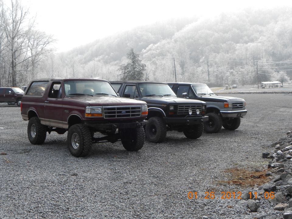 Maroon Bronco and two dark trucks parked on gravel lot with misty hills in background