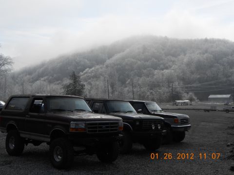 1986 Ford Bronco - Three Ford Broncos are parked in a lot with a misty mountain range behind them.