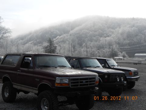 1986 Ford Bronco - Three Ford Broncos are parked in a line on a foggy day, with a mountain range in the background.